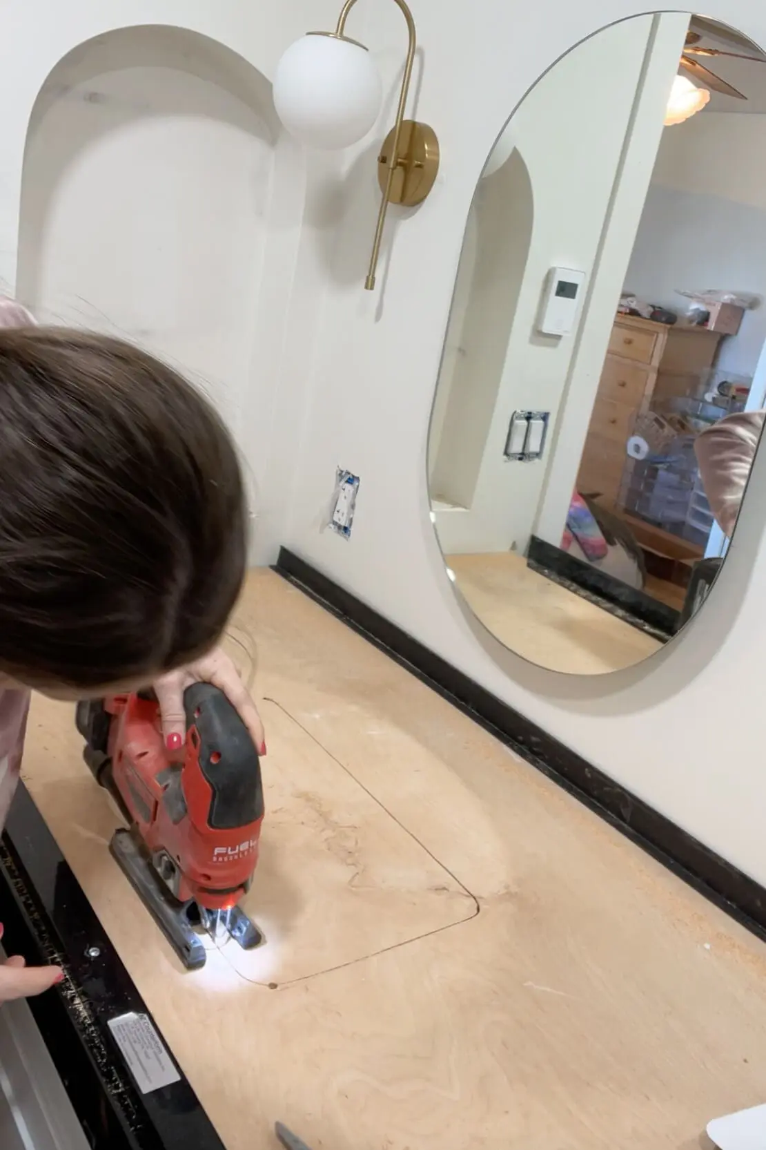 A woman using a red jigsaw to cut a rectangular outline into a wooden countertop base for a concrete countertop installation. The workspace features a black plastic edge frame, a gold wall sconce, and an oval mirror on the wall.